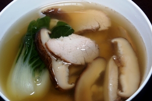 a white bowl containing Chinese chicken broth, wontons, bok choy, barbecued pork and Chinese black mushrooms